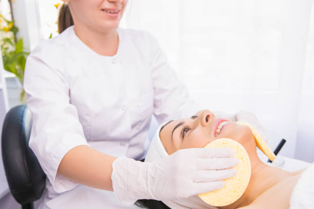 istock Professional cosmetologist making cleansing procedure for moisturizing the face with two sponges to a woman in a beauty salon. 1352912565