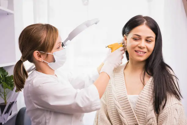 Specialist in medical mask and gloves pierces the client's ears with a piercing gun in the cosmetologist cabinet. High quality photo