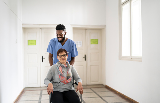 A smiling male healthcare worker pushes a senior white female patient along a light bright white corridor in a hospital