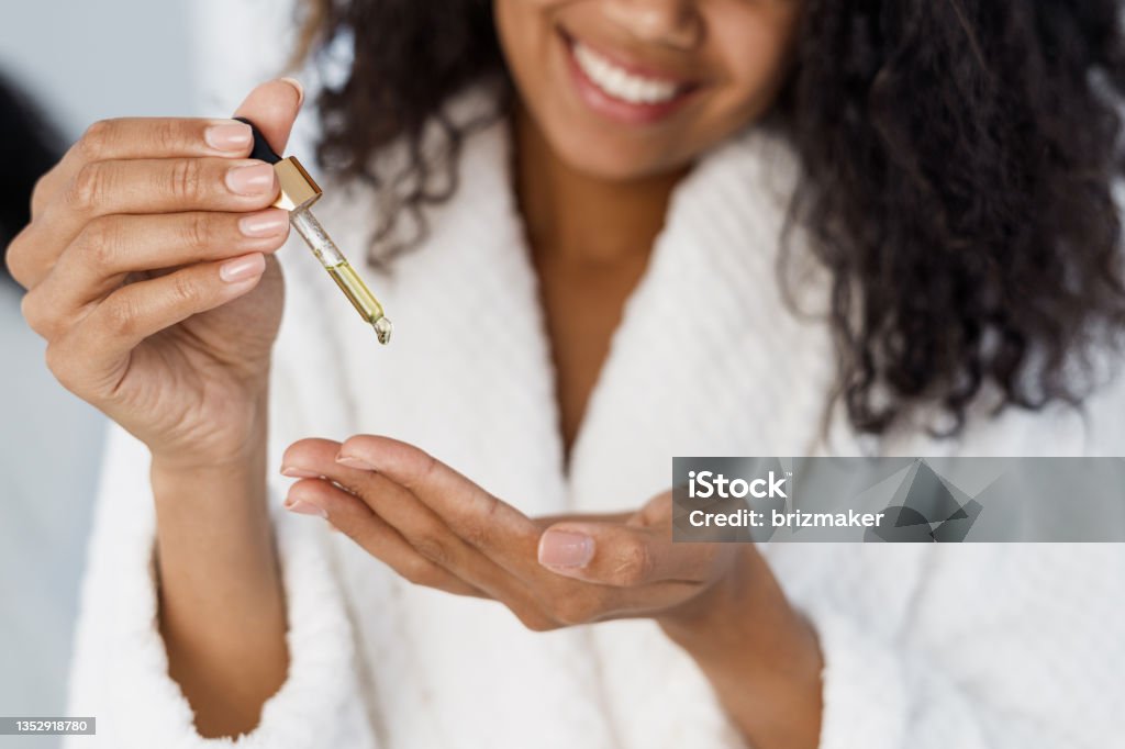 Young african american woman holding pipette with serum oil Nail care advertising concept. Cropped view of young happy afro american woman in bathrobe holding pipette with cuticle oil in hand, applying serum on fingers, close up Essential Oil Stock Photo