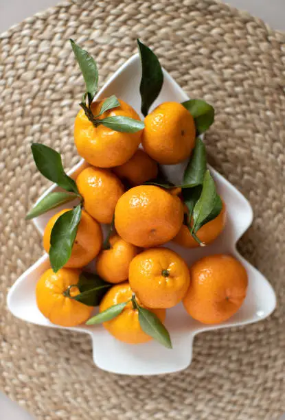 Photo of Small ripe tangerines with leaves, on plate in shape of Christmas tree on wicker serving napkin. Concept winter vitamin food. Moody still life festive composition. For greeting card, book, stories