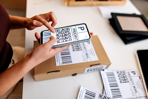 Female warehouse worker, seller, drop shipping small business owner holding phone scanning retail package postal parcel bar code on e commerce shipping box label on smartphone using mobile app, close up