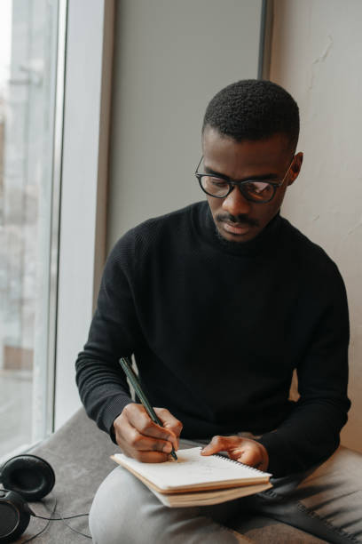 istock Busy black man sitting on a windowsill, working, writing notes in a notepad 1352927522