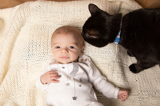 A happy 2.5 month old baby boy looks at the camera as a black cat steps in to sniff him.