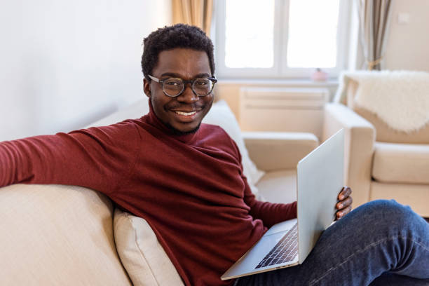 portrait d’un homme afro-américain souriant avec des lunettes assis sur un canapé au bureau travaillant sur un ordinateur portable - looking at camera smiling desk isolated photos et images de collection