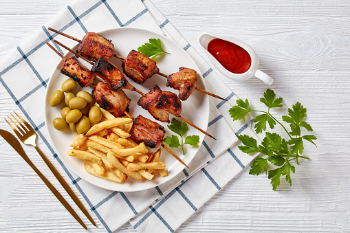 Juicy grilled pork kebabs on a plate with green olives and potato fries, horizontal view from above, flat lay, free space