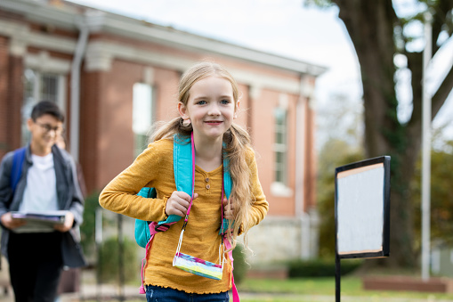 A sweet little girl with pigtails giggles outside as she poses for a portrait in front of her school.  She is dressed casually and is holding her books tightly as her peers play away in the background.