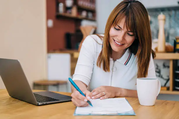 Shot of an adorable smiling businesswoman using laptop and making notes on a clipboard at home.
