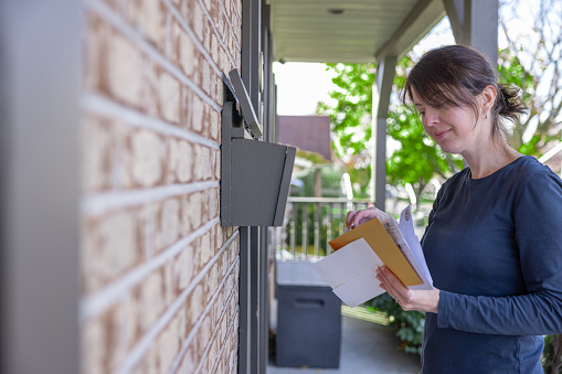 A woman is collecting post at home in her mailbox in Australia. She is smiling and  picking-up her mail. She is looking at the letters she received.
