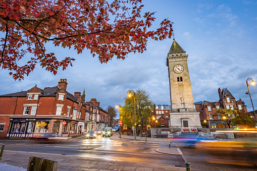 Identifiable stores and cars are visible in this image. The war memorial clock tower in Leek with visible names as well as other stores and car number plates. The memorial was commissioned by Sir Arthur Nicholson and his wife when their son died in Ypres, Belgium, during World War 1 and opened on 20 August 1925. It was intended as a memorial to all local men who died in that war and later to the dead of World War 2 as well. Made of Portland stone, it's one of the tallest war memorials in the UK.