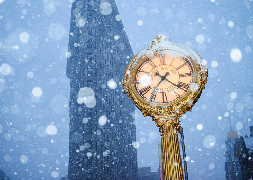 Antique Fifth Avenue Clock covered with snow, Flatiron Building on the background, New York, USA