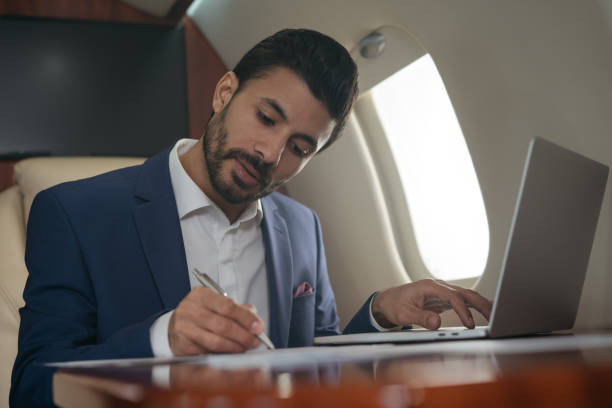 бизнесмен пользуется ноутбуком, печатает, работает онлайн сидя в самолете. уверенный ближневосточный предприниматель делает заметки, подп - only men mid adult men men author стоковые фото и изображения