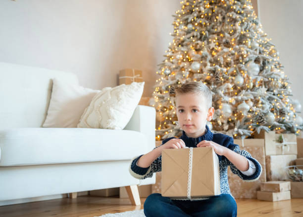 小さな子供の男の子は、プレゼントのxmasを開梱します。ギフトボックスとクリスマスツリーの近くにかわいいブロンドの髪の子供。装飾されたリビングルーム。魔法の時間。大晦日。 - gift little boys people blond hair ストックフォトと画像