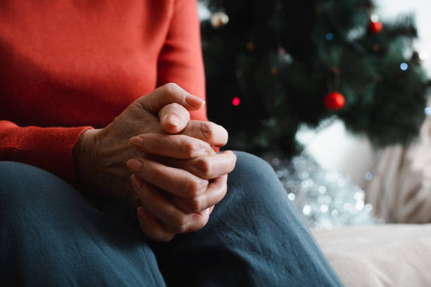 Lonely senior woman sitting at home in Christmas celebration. Close-up of an elderly woman's hand against background of decorated Christmas tree. Loneliness, sad holiday concept stock photo
