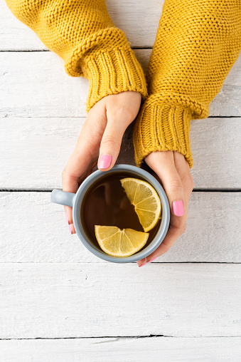 Overhead shot of woman’s hands warm sweater holding mug of tea with lemon on white wooden background with copyspace