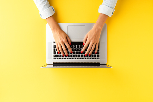 Woman’s hands working on laptop. Modern office desktop with accessories on yellow background with copyspace. Top view