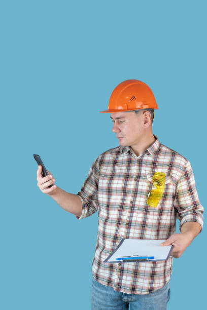 istock A worker in an orange helmet reads text messages while holding a mobile phone and clipboard with papers on a blue background 1353021495