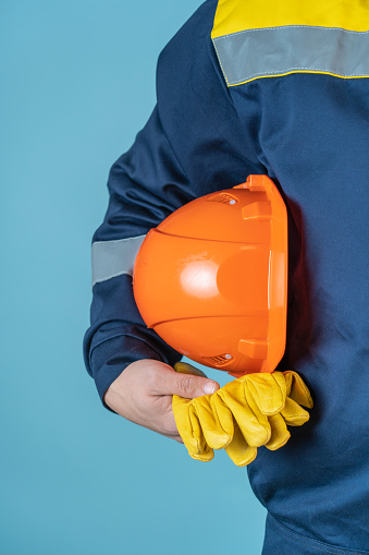 A worker in overalls holds an orange protective helmet and yellow gloves on a blue background in his hand. The concept of safe work on the construction site.