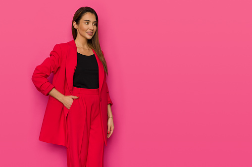 Beautiful young woman in elegant red jacket and baggy pants is posing with hand in pocket and looking away. Studio shot on pink background.