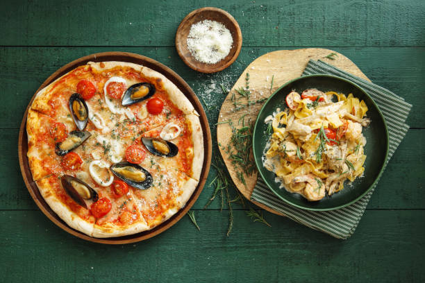 Pizza with shrimps, squids, scallops, mussels and capers. Italian fettuccine with prawns, salmon and herbs. Flat lay top-down composition on dark green background.