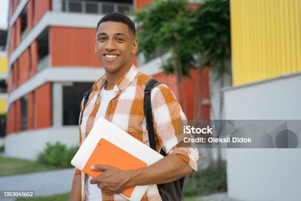 Confident Developer Holding Book And Laptop Walking On Urban Street Handsome Smiling African American Student Student Looking At Camera In University Campus Education Concept Stock Photo - Download Image Now