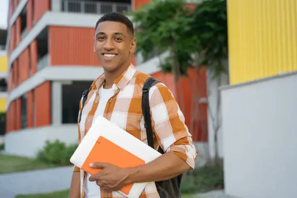 Photo of Confident developer holding book and laptop walking on urban street. Handsome smiling African American student student looking at camera in university campus. Education concept