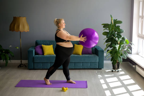 Pregnant lady is doing Pilates at home, lifting a fit ball above herself. A fat, obese pregnant lady is doing Pilates at home, lifting a fit ball above herself. Home workouts in Interior room with a large window, sofa and yellow-purple pillows. Weight loss in quarantine fitness ball photos stock pictures, royalty-free photos & images