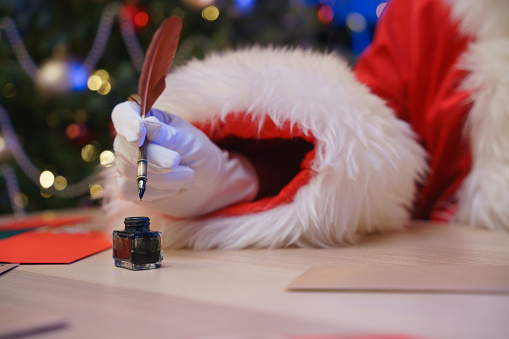 Close-up. Santa’s hand is dipping a pen in an ink pot. A Christmas tree decorated with balls, lights and beads next in the background. Colorful envelops are on a table.