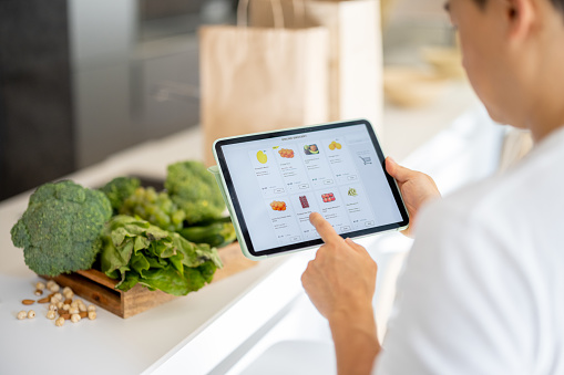 Man watching groceries in online store on digital tablet. Concept of online shopping and e-commerce. Idea of healthy and vegetarian eating. Person on home kitchen with table full of organic products