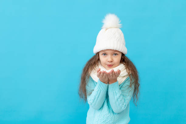 urocza dziewczynka w zimowym ubraniu czapka i sweter zdmuchuje śnieg z rąk na niebieskim izolowanym tle, miejscu lub miejscu na tekst - propeller beanie zdjęcia i obrazy z banku zdjęć
