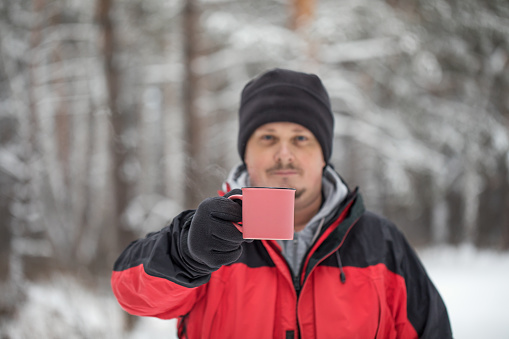 A mug with a hot drink in the hands of a man. Portrait of a man for a walk in the winter forest.