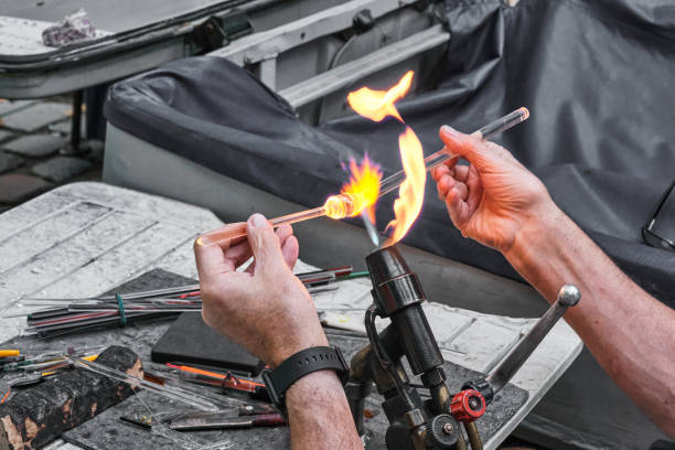 Hands of mature glassblower holding glass rod over burner flame. Process of artistic glass processing, flamework. Hands of mature glassblower holding glass rod over burner flame over background of workplace and tools. Process of artistic glass processing, lampwork. Craft concept hand blown glass stock pictures, royalty-free photos & images