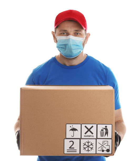 istock Courier in mask holding cardboard box with different packaging symbols on white background. Parcel delivery 1353097818