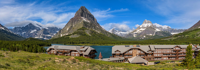 Many Glacier, Montana, USA - June 11, 2021: A panoramic view of Swiss chalet style historic Many Glacier Hotel at shore of Swiftcurrent Lake, with rugged snow peaks in background, on a sunny Spring morning  in Glacier National Park.
