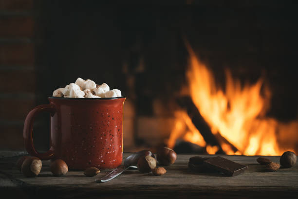 kakao mit marshmallows und schokolade in einem roten becher auf einem holztisch in der nähe eines brennenden kamins - behaglich stock-fotos und bilder