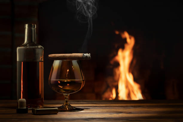 焼け付いた暖炉の近くのテーブルの上にコニャック、葉巻、ボトルのグラス。リラクゼーションと楽しいコンセプト - brandy snifter ストックフォトと画像