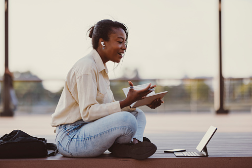 Happy African-American woman using digital tablet and bluetooth earphones for E-Learning while holding notebook and sitting on wooden bench