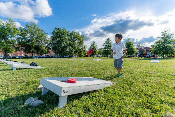 A young boy is tossing bean bags at a corn hole board during a summer afternoon with some clouds in the sky.