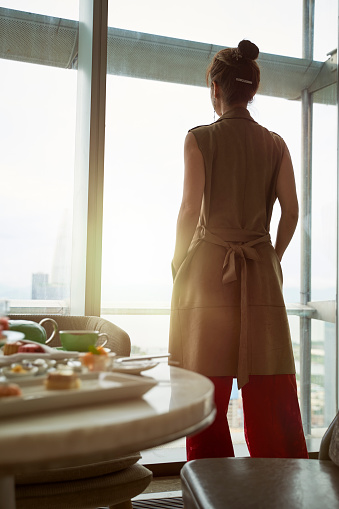 mature asian woman looking at view through window of hotel room with breakfast on table