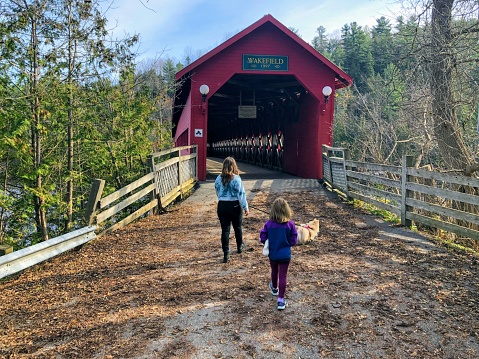 A mother and daughter walking together through the red Wakefield covered bridge along the Gatineau River on a beautiful sunny fall day in pretty town of Wakefield, Quebec, Canada.