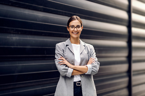 Una joven y exitosa empresaria de pie frente a un fondo gris con los brazos cruzados y mirando a la cámara. Una mujer de negocios posando afuera photo