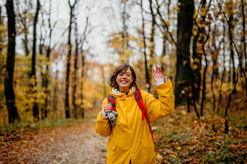 In this image a cheerful young adult in a yellow raincoat is waving at the camera while smiling in an autumn forest environment