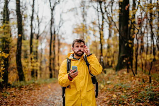A handsome man with a beard wearing a yellow raincoat scratches his head as he looks around while holding his phone