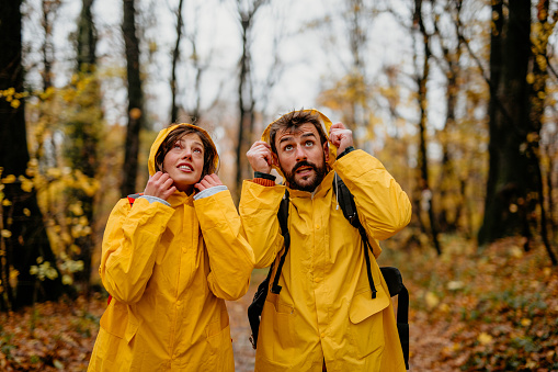 Portrait of two young travelers in raincoats, putting a hood on the head and checking the weather while walking through the forest.