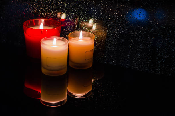 Blurred focus of candles in glass put beside window that have rain drop in dark background.