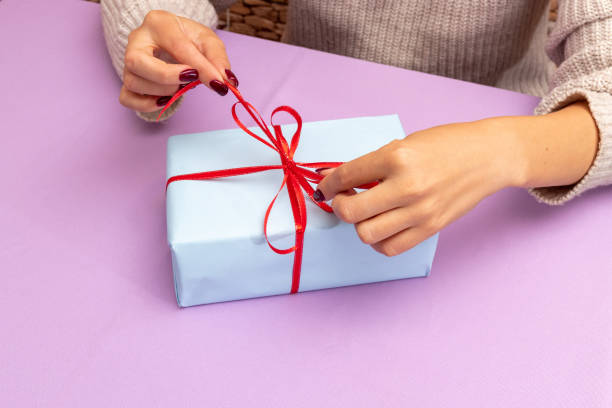 Women's hands are pulling red bow on gift. Purple background with place for text. Advertising of gifts. Selective focus. stock photo
