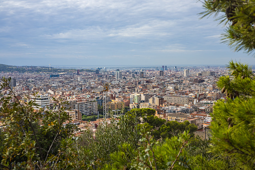 Barcelona, Spain - September 22, 2021: View through the trees from Park Guell over the city. Aerial View Cityscape with cloudy sky. Surrounded by trees with green leaves