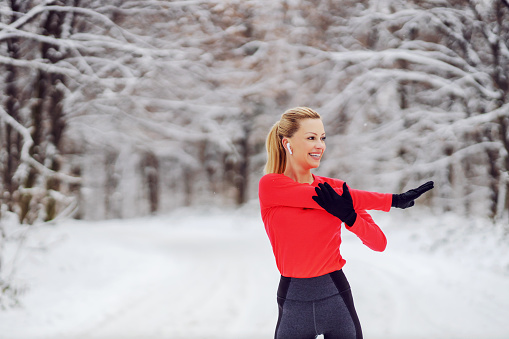 Fit sportswoman standing in nature and doing warmup and stretching exercises at snowy winter day. Sportswear, exercises in nature, winter fitness