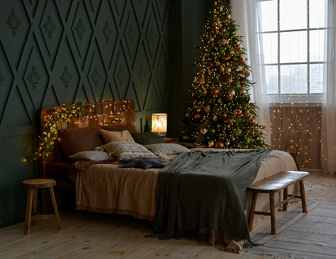 Interior of a beautiful Christmas bedroom with a wooden bed, bench, ,beautifully decorated Christmas tree by the window. Christmas decorations. Happy New Year and Merry Christmas!