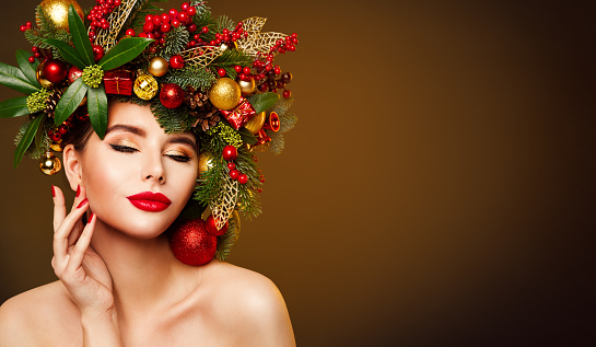 Christmas Model Beauty in Decorated Fir Tree Wreath. Woman Face and Body Skin Care. Girl with Red Lips Make up Touching Cheek over Dark Studio Brown Background with Copy Space